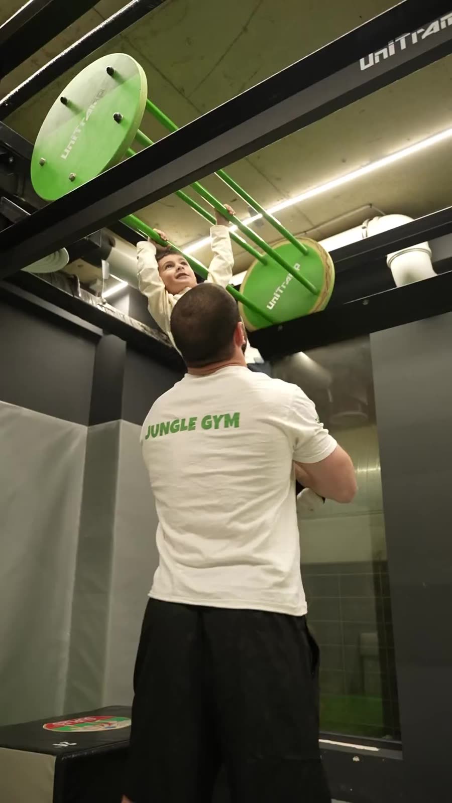 Gymnasia coach in a JUNGLE GYM shirt lifting a young child to reach the green climbing bars.
